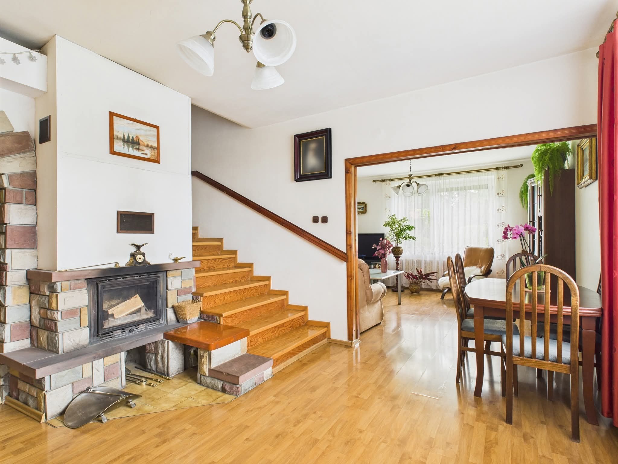 Jasne wnętrze domu z kominkiem obłożonym kamieniem, drewnianymi schodami i parkietem. Widok na salon przez otwarte drzwi. Ciepłe oświetlenie.