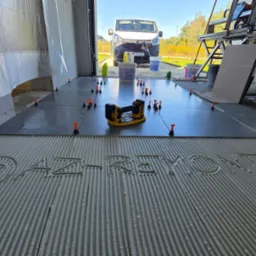 Układanie ciemnych płytek na podłodze garażu z użyciem poziomowania. Na kleju napisana nazwa firmy. W tle samochód i narzędzia glazurnicze.