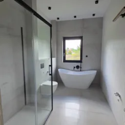 Jasna łazienka z kabiną prysznicową, wolnostojącą wanną i podwieszaną toaletą. Minimalistyczny design z czarnymi akcentami oświetlenia i armatury.