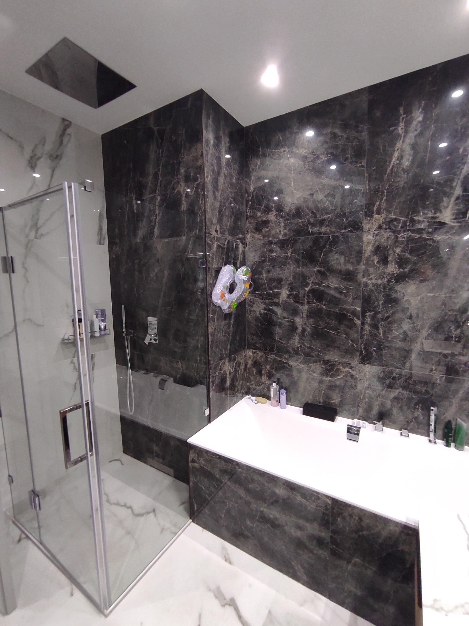 Nowoczesna łazienka z kabiną prysznicową ze szklanymi drzwiami i ścianami wyłożonymi ciemnym marmurem, biała wanna z czarnym akcentem, na ścianie zawieszona zabawka w kształcie pistoletu na wodę.