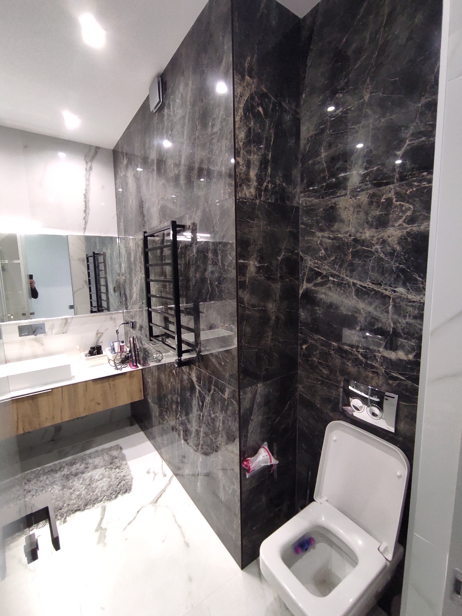 Nowoczesna łazienka z marmurowymi płytkami w odcieniach szarości i czerni, wiszącą toaletą z chromowanym przyciskiem spłukiwania, drewnianą szafką pod umywalką i grzejnikiem drabinkowym.