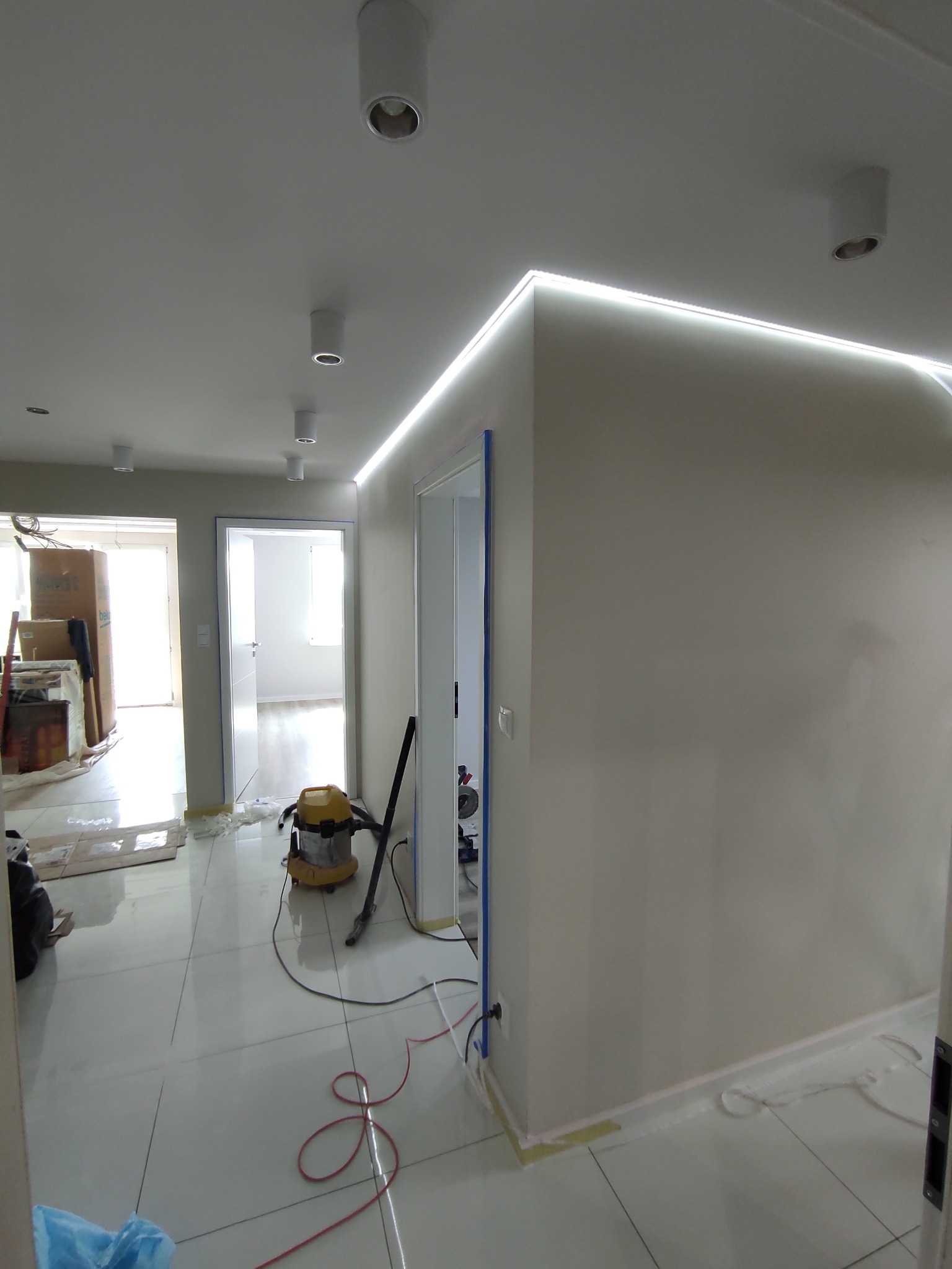 Wnętrze w trakcie prac wykończeniowych: białe płytki podłogowe, świecąca listwa LED pod sufitem, odkurzacz przemysłowy i elementy zabezpieczające krawędzie. Widoczne gniazdko elektryczne.
