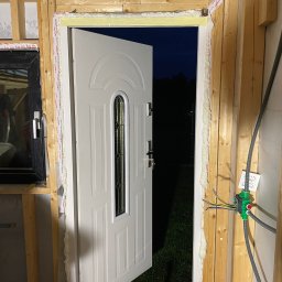 Shomnikov Pavlo - Białe drzwi wejściowe z szybą, częściowo otwarte, zamontowane w surowym otworze z drewnianą ramą, uszczelnione pianką montażową; widoczny fragment instalacji elektrycznej i gniazdko.