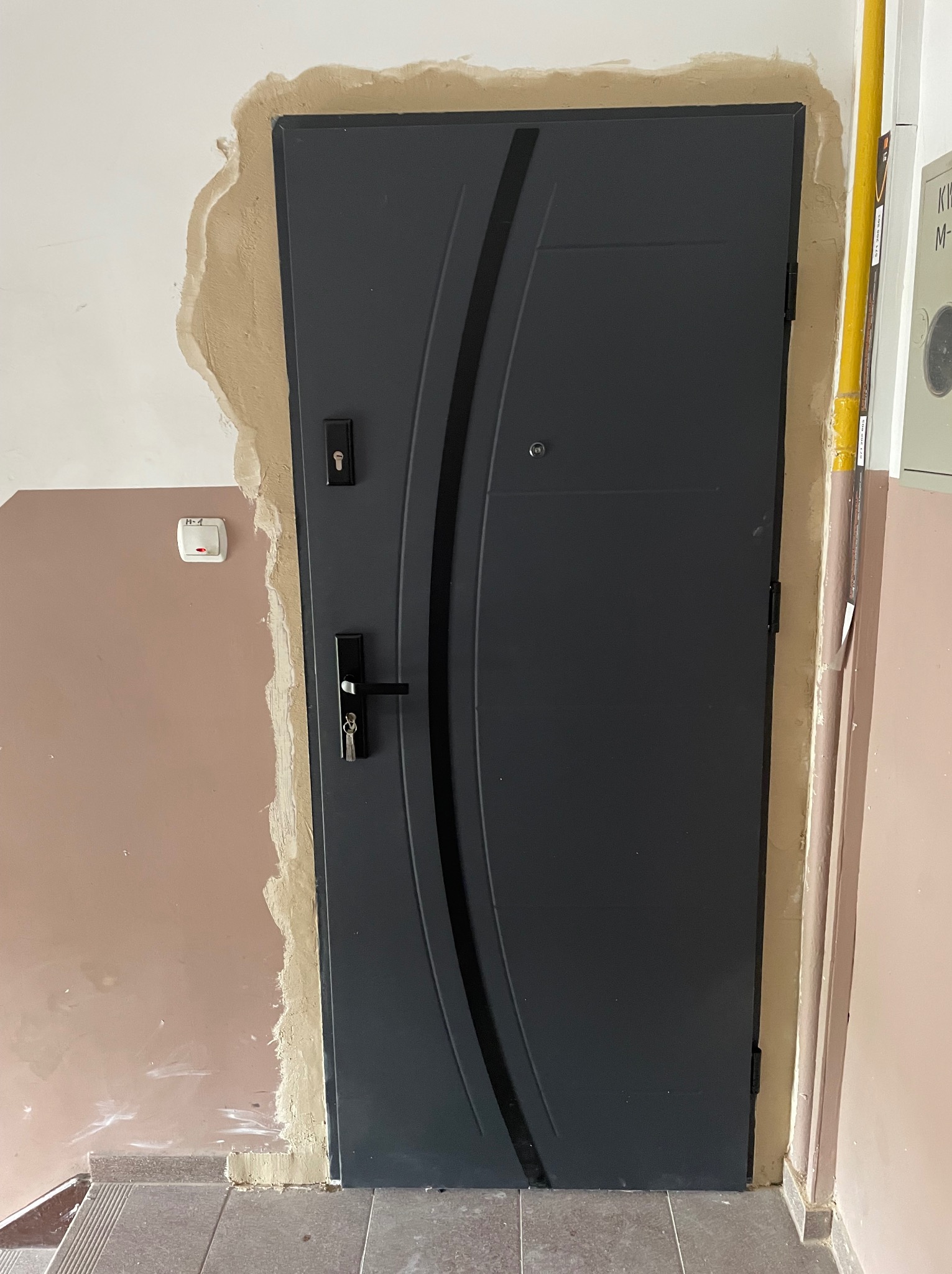 Szare drzwi wejściowe z czarną klamką i wkładką, widoczne ślady po montażu na ścianie, zarysowane ściany i brakujące wykończenie wokół ościeżnicy.