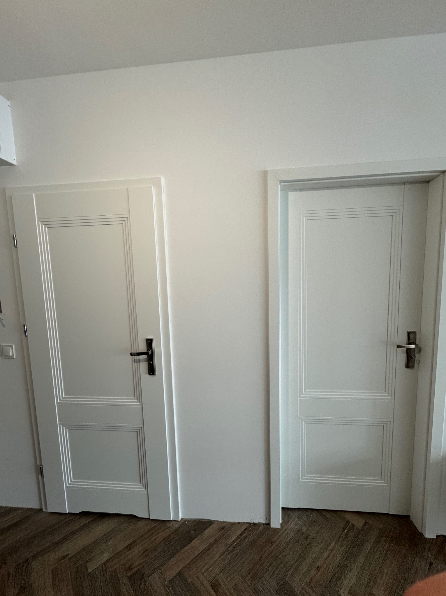 Dwa białe, panelowe drzwi wewnętrzne z czarną klamką po lewej i srebrną po prawej, zamontowane w białej ścianie nad parkietem ułożonym w jodełkę.