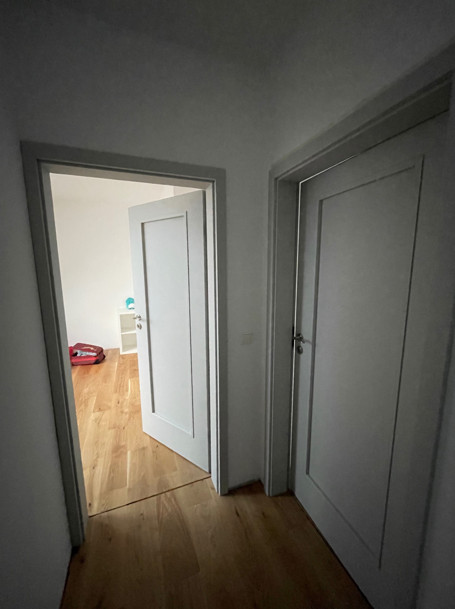 Dwa białe drzwi w korytarzu, jedne otwarte do pokoju z drewnianą podłogą, gdzie widać walizkę i białą szafkę.