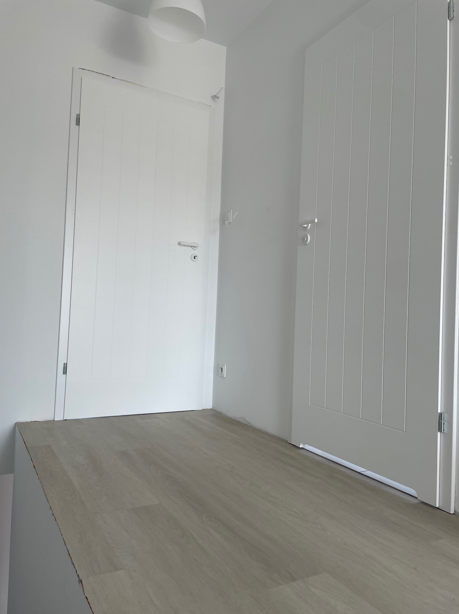 Dwie białe, wewnętrzne drzwi z pionowymi frezowaniami w minimalistycznym, jasnym wnętrzu. Jedne drzwi zamknięte, drugie otwarte, ukazujące fragment korytarza z panelami podłogowymi.