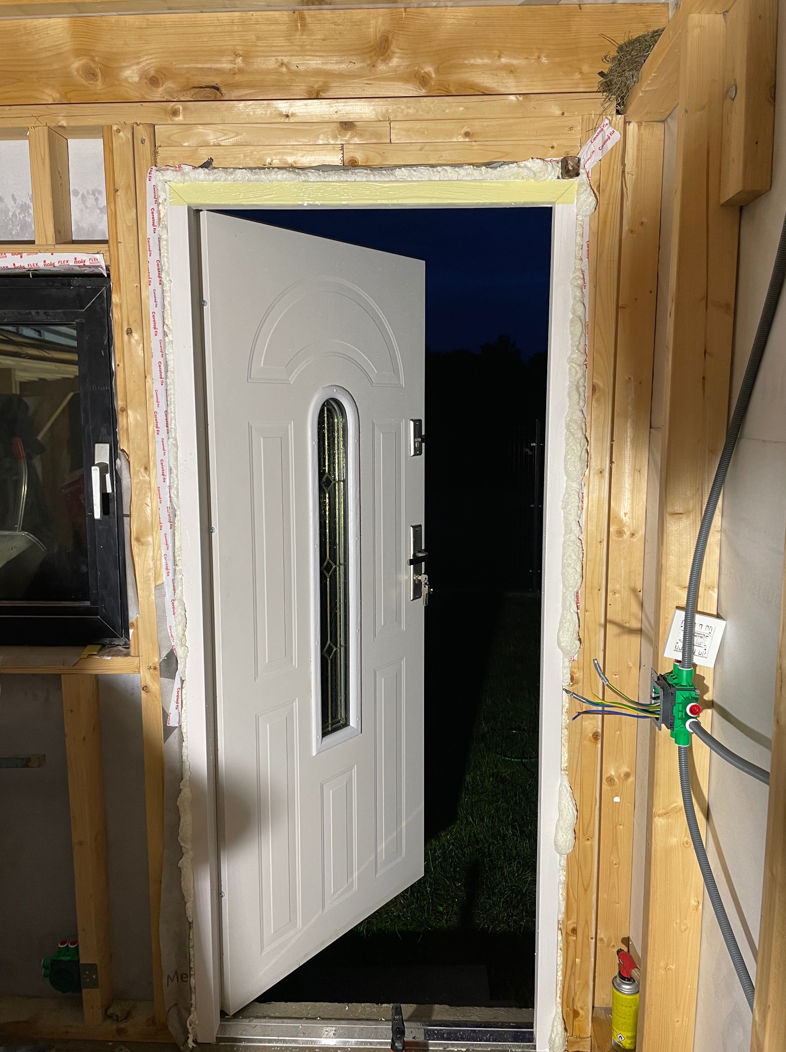 Białe drzwi wejściowe z szybą, częściowo otwarte, zamontowane w surowym otworze z drewnianą ramą, uszczelnione pianką montażową; widoczny fragment instalacji elektrycznej i gniazdko.