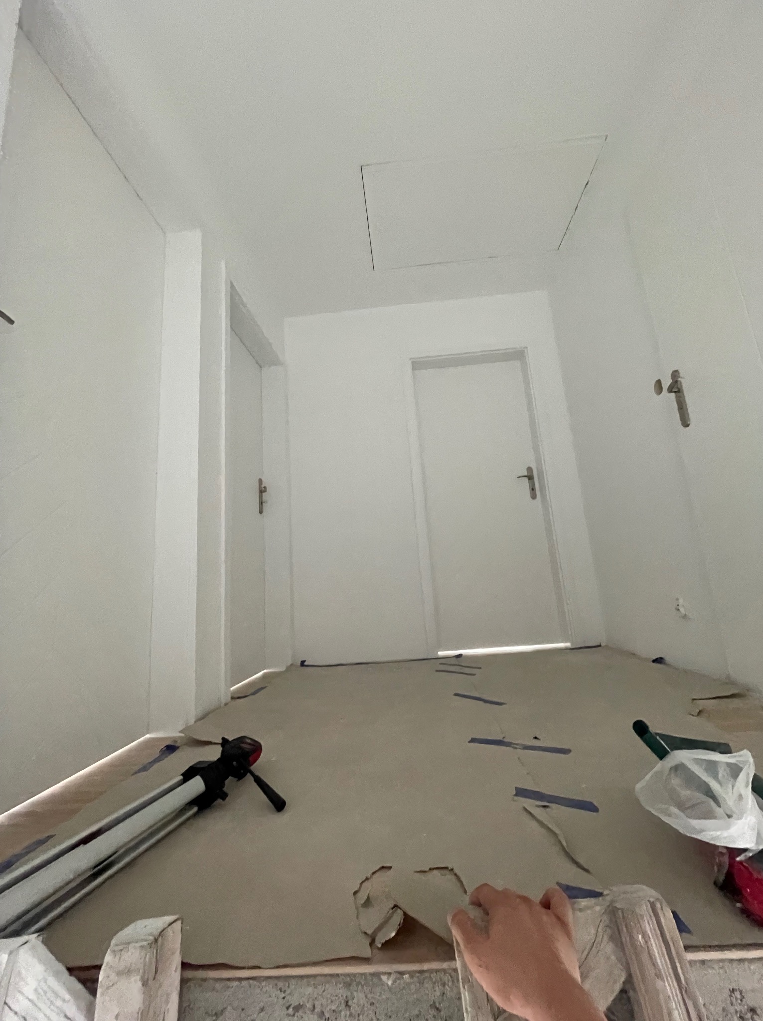 Korytarz w trakcie remontu z zabezpieczoną podłogą, widoczne dwie białe drzwi i narzędzia, fragment dłoni w kadrze.