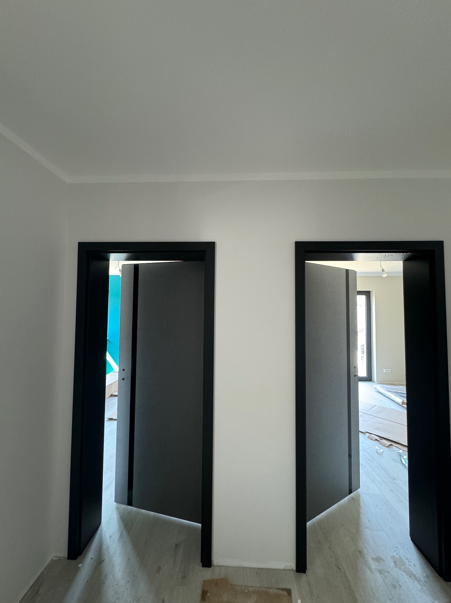Dwa otwarte, nowoczesne drzwi wewnętrzne z czarnymi ościeżnicami i szarymi skrzydłami, widoczne w korytarzu z jasnymi ścianami i podłogą w trakcie wykończenia.