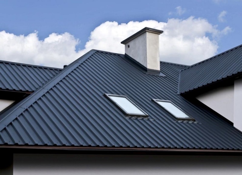 Grafitowy dach z blachy trapezowej z dwoma oknami dachowymi i kominem na tle błękitnego nieba z lekkimi obłokami.
