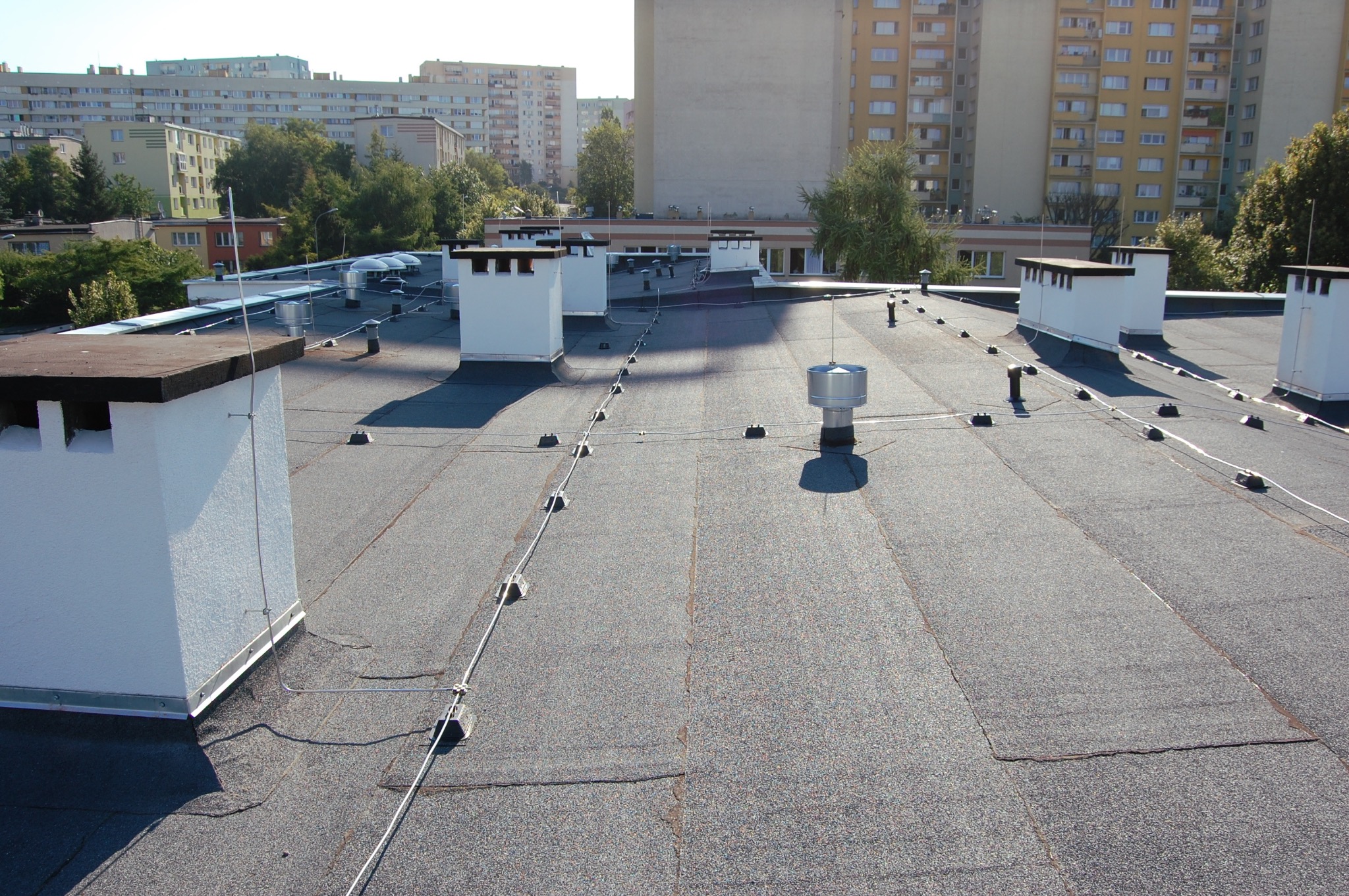 Płaski dach pokryty papą termozgrzewalną z kominami wentylacyjnymi i systemem odgromowym, widok z góry na tle zabudowy miejskiej.