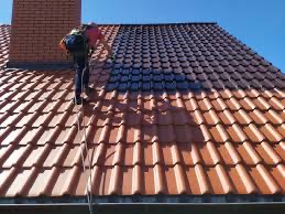 Pracownik w uprzęży asekuracyjnej czyści dach z czerwonej dachówki ceramicznej, widoczny fragment dachu po czyszczeniu, jaśniejsza i ciemniejsza część, błękitne niebo w tle.