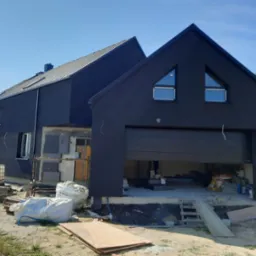 Nowoczesny dom z ciemną elewacją w trakcie prac wykończeniowych, widoczne materiały budowlane i otwarty garaż.