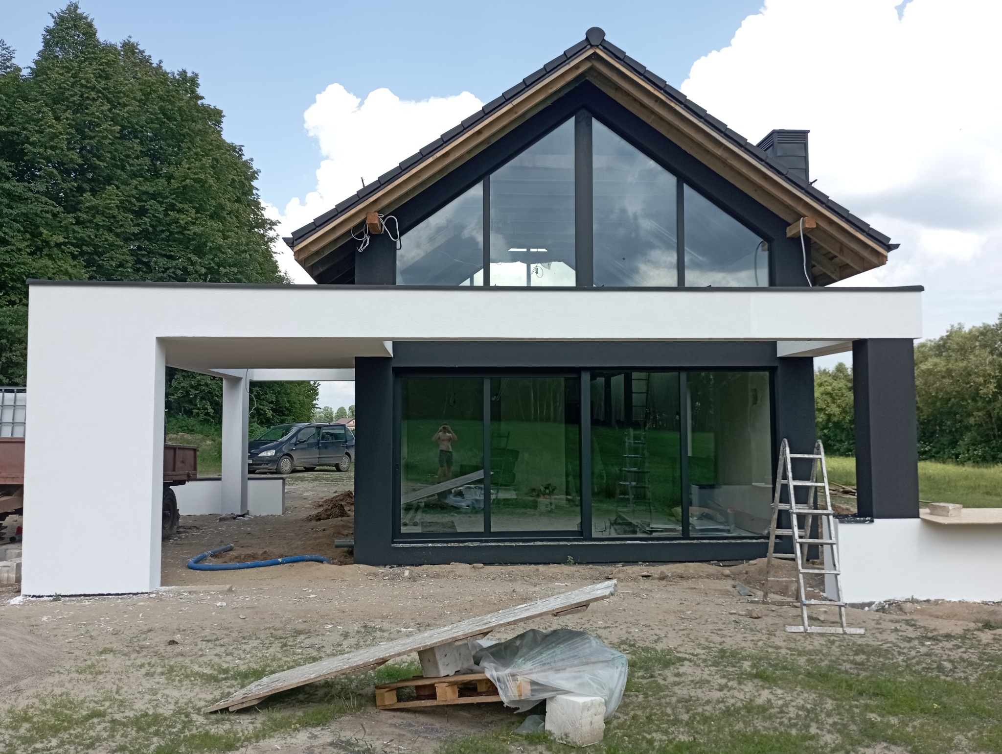 Nowoczesny dom jednorodzinny w trakcie wykańczania elewacji, widoczne białe i grafitowe fragmenty ścian, duże okna odbijające otoczenie, drabina oparta o ścianę, teren wokół domu w trakcie prac...