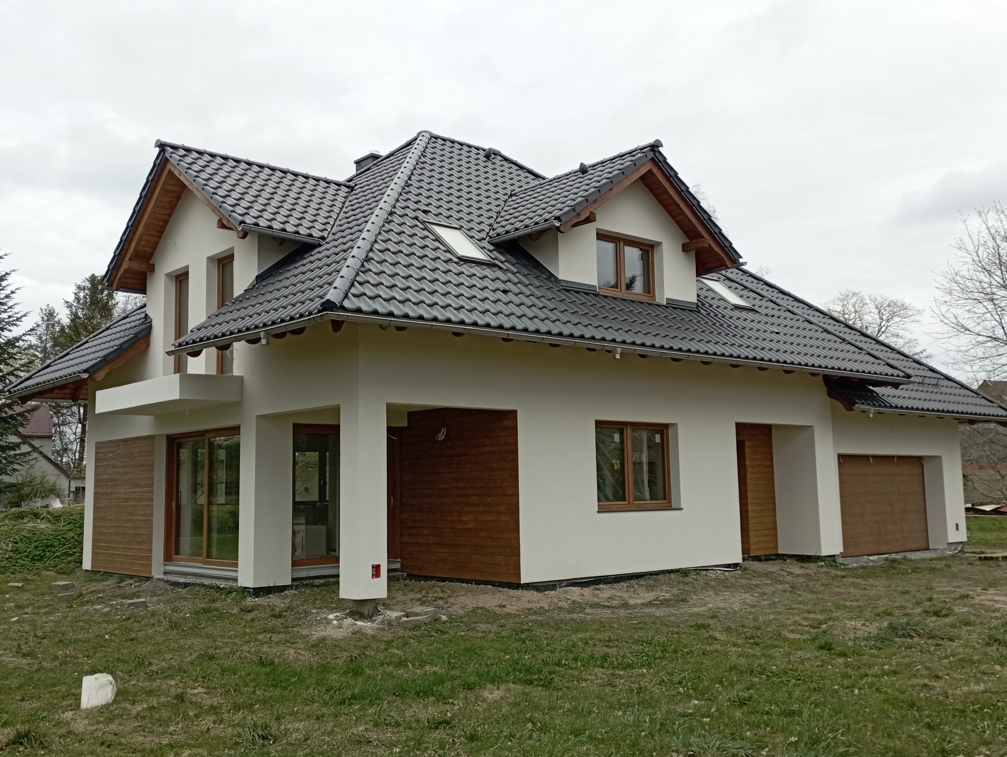 Nowoczesny dom jednorodzinny z białą elewacją, drewnianymi akcentami i czarną dachówką, widok z zewnątrz w pochmurny dzień.