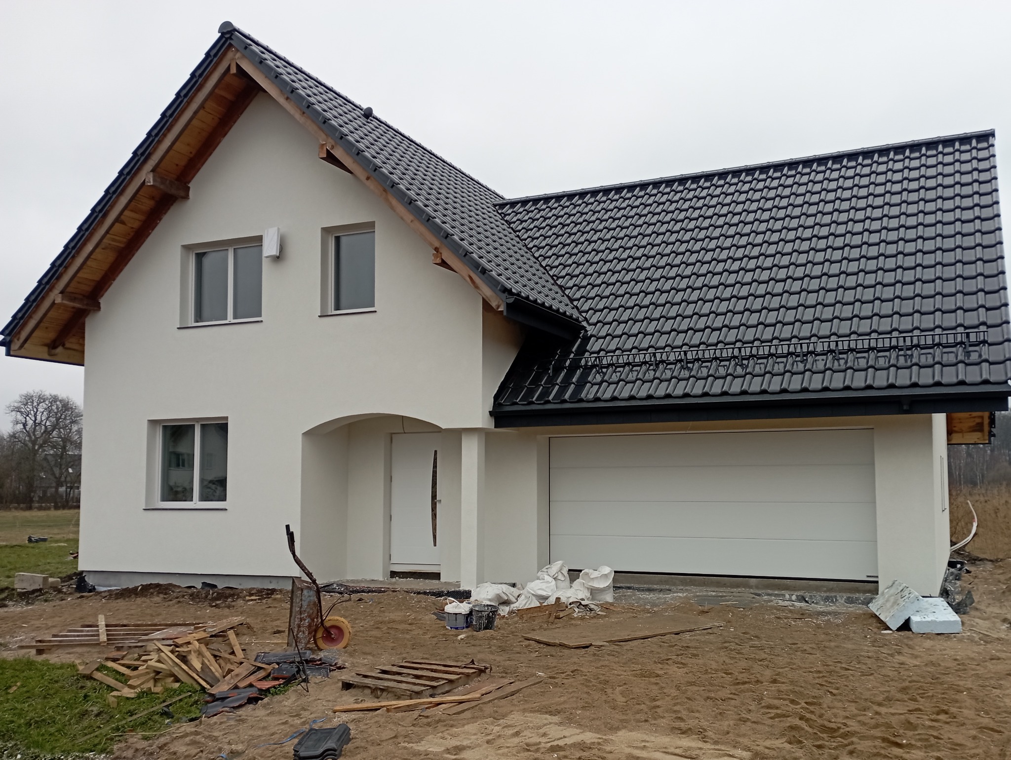 Nowoczesny dom jednorodzinny z białą elewacją, czarną dachówką i garażem, widok z zewnątrz, teren wokół domu w trakcie prac wykończeniowych, widoczne materiały budowlane i taczka.