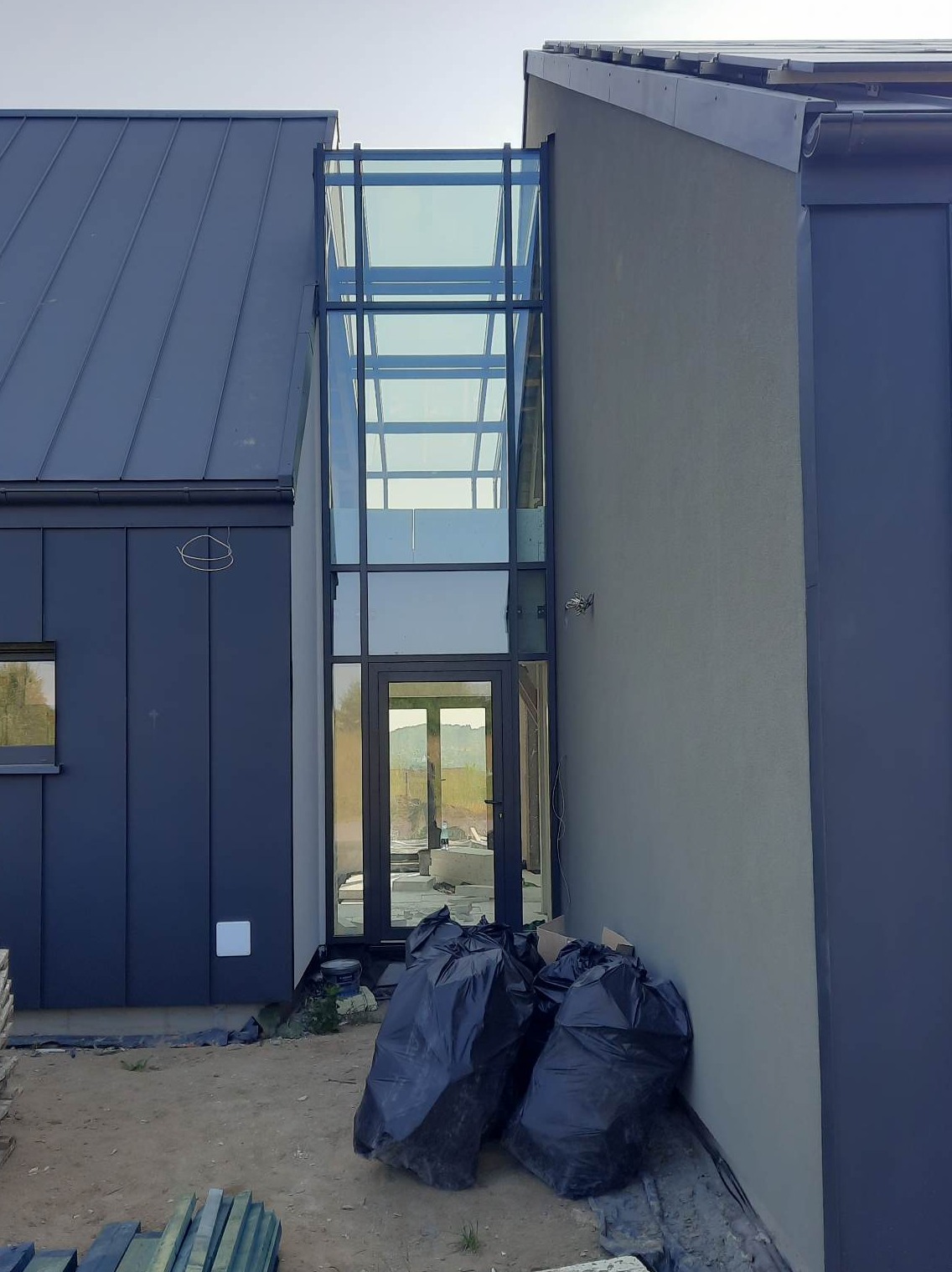 Nowoczesny dom z przeszkloną klatką schodową, widoczne worki z odpadami budowlanymi i materiały przygotowane do dalszych prac wykończeniowych.