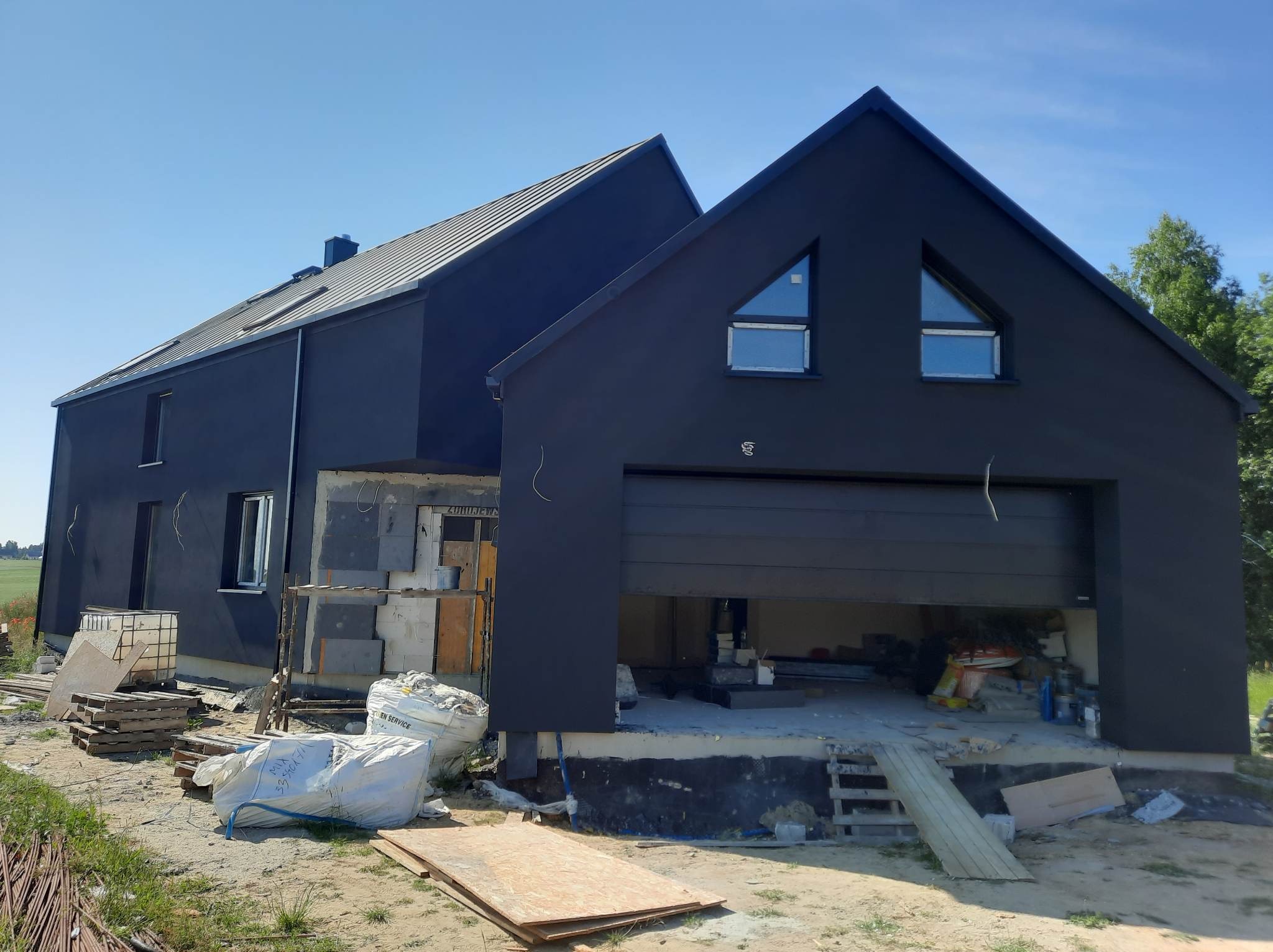 Nowoczesny dom z ciemną elewacją w trakcie prac wykończeniowych, widoczne materiały budowlane i otwarty garaż.