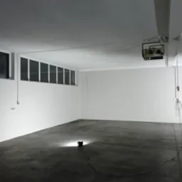 Puste, białe pomieszczenie biurowe z betonową podłogą, oświetlone pojedynczą lampą podłogową, widoczne okna pod sufitem i instalacja wentylacyjna pod stropem.