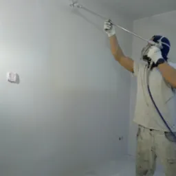 Pracownik w kombinezonie ochronnym maluje ścianę natryskowo, widoczny fragment zabezpieczonej ściany i rozpylona farba w powietrzu.