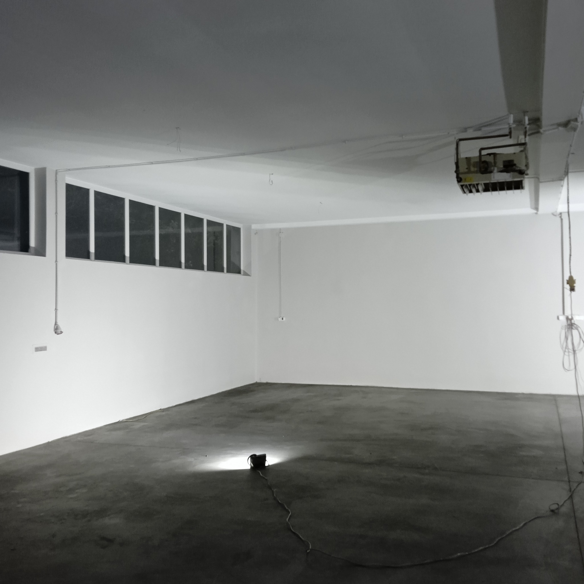 Puste, białe pomieszczenie biurowe z betonową podłogą, oświetlone pojedynczą lampą podłogową, widoczne okna pod sufitem i instalacja wentylacyjna pod stropem.