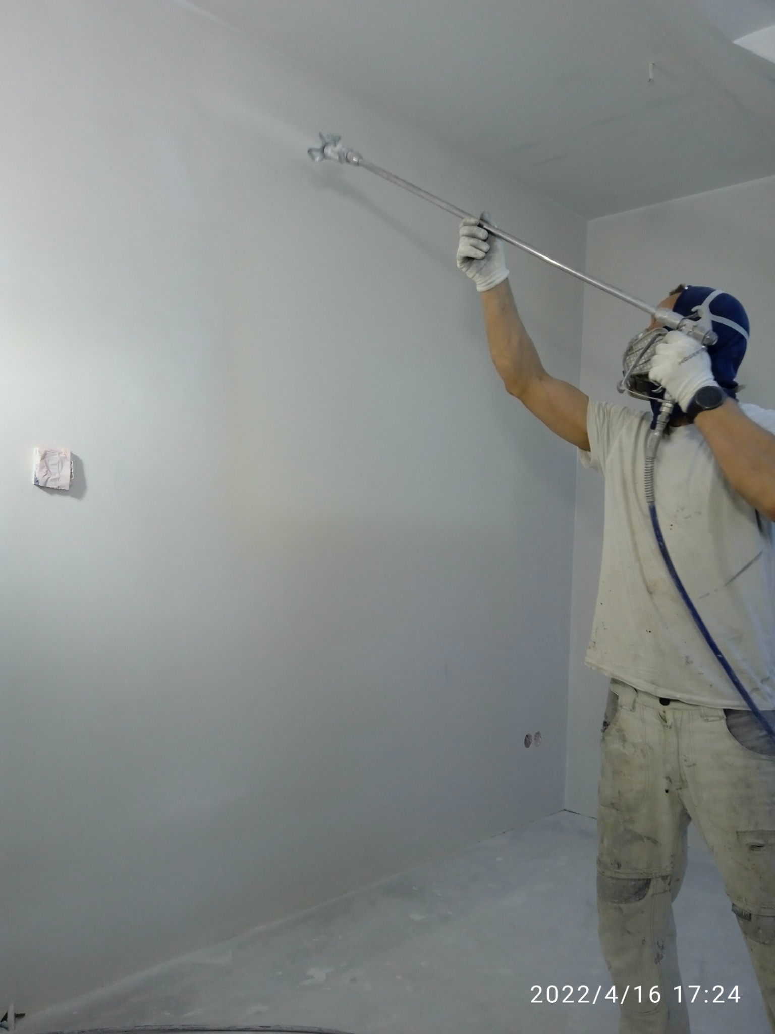 Pracownik w kombinezonie ochronnym maluje ścianę natryskowo, widoczny fragment zabezpieczonej ściany i rozpylona farba w powietrzu.