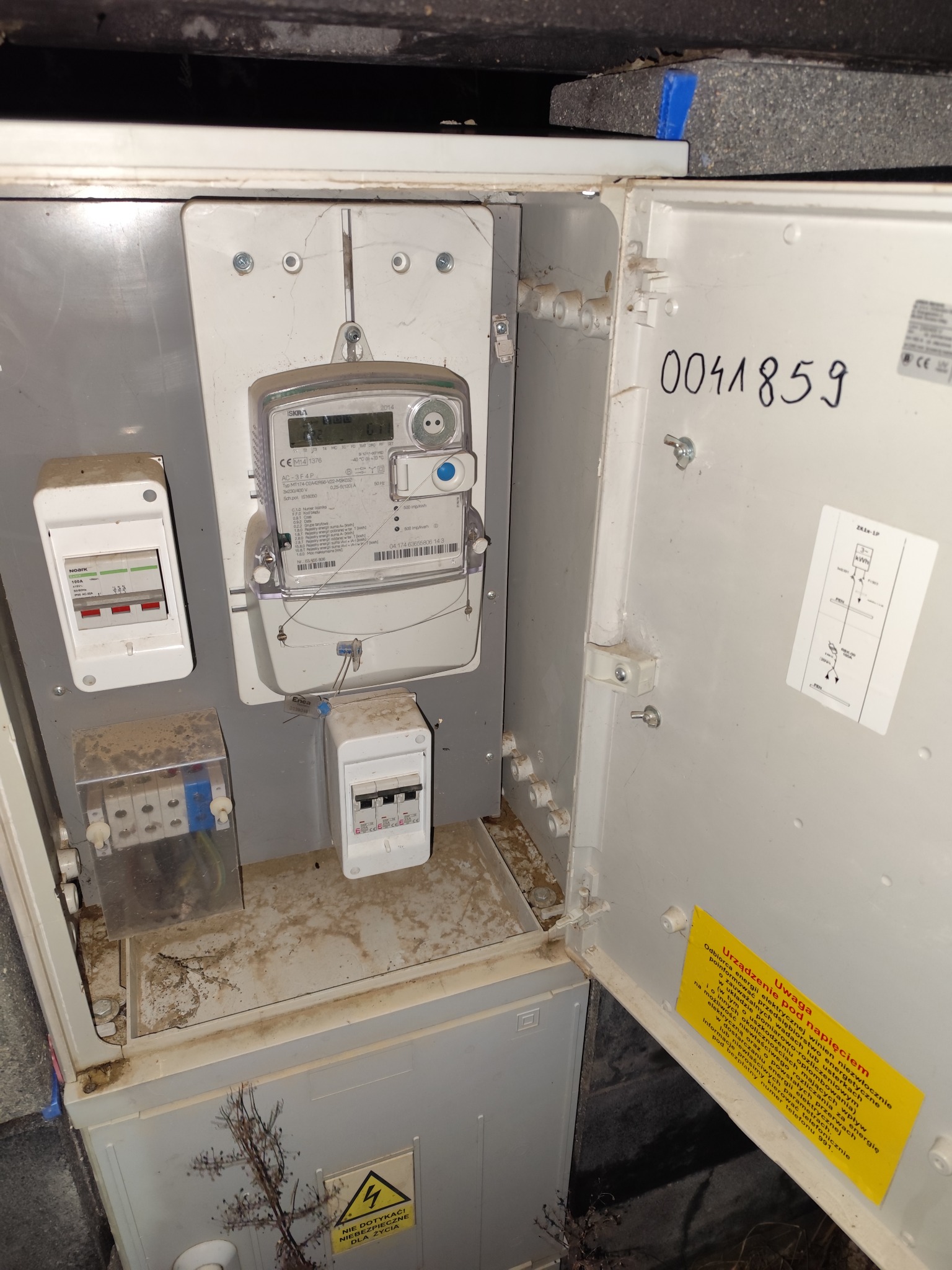 Otwarta skrzynka elektryczna z licznikiem energii, bezpiecznikami i ostrzeżeniem o napięciu, widoczne zabrudzenia i kurz na elementach.