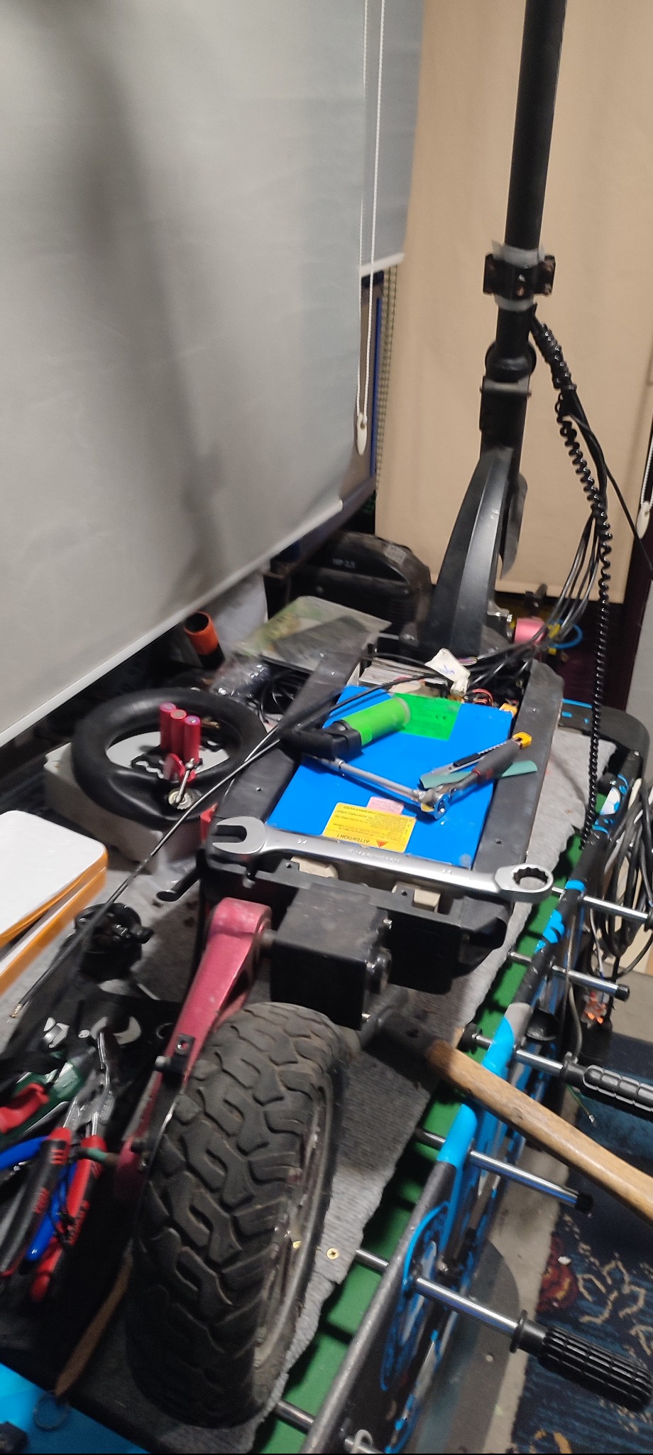 Elektryczna hulajnoga w trakcie naprawy, widoczne narzędzia, akumulator i części zamienne na stole warsztatowym w garażu.