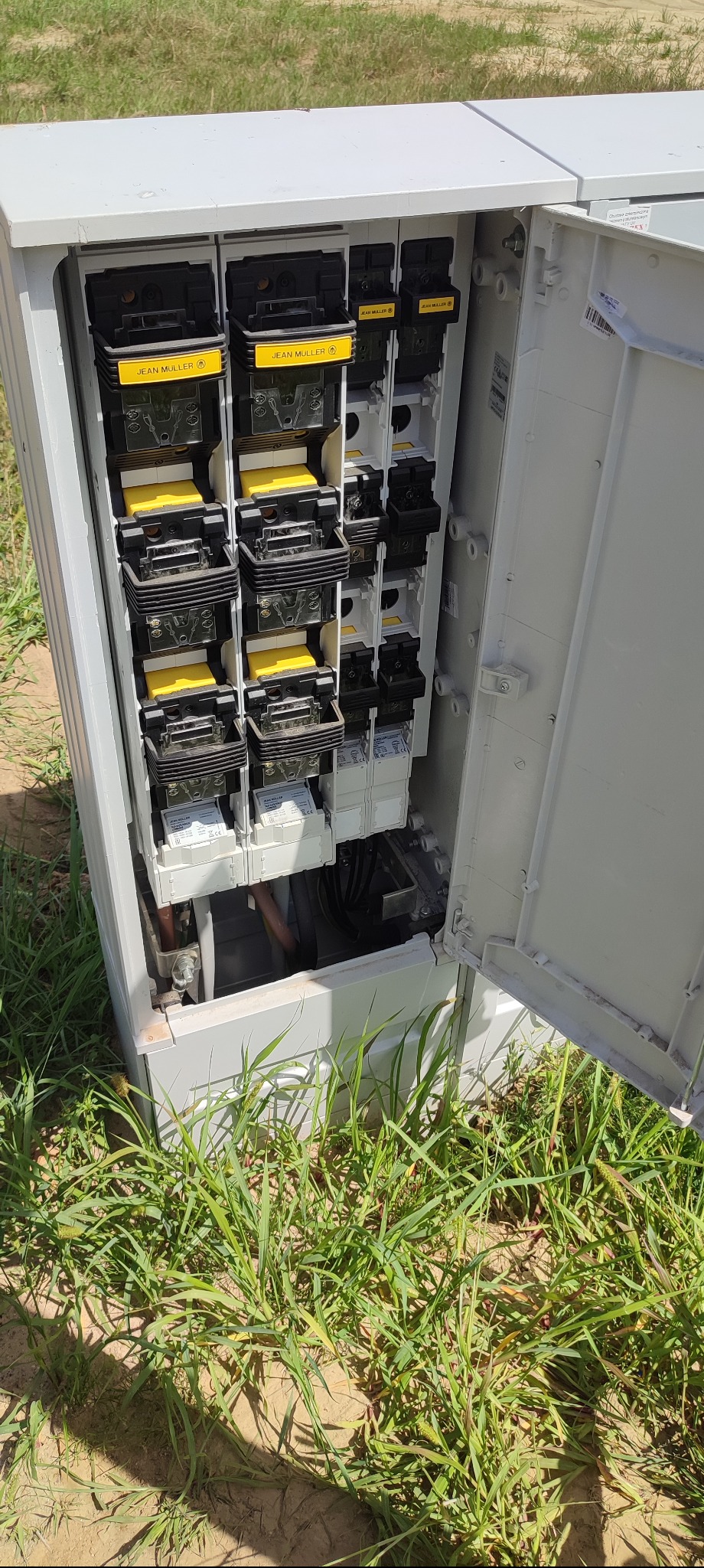 Otwarta skrzynka elektryczna z bezpiecznikami Jean Muller na zewnątrz, widoczne kable i elementy instalacji, otoczona trawą.
