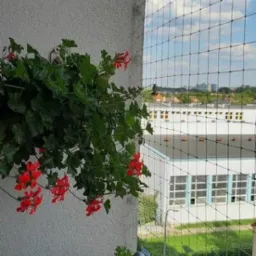 Zamontowana siatka ochronna na balkonie, zabezpieczająca przed gołębiami, widok z bliska z kwiatami w doniczce na pierwszym planie, w tle budynki i błękitne niebo.