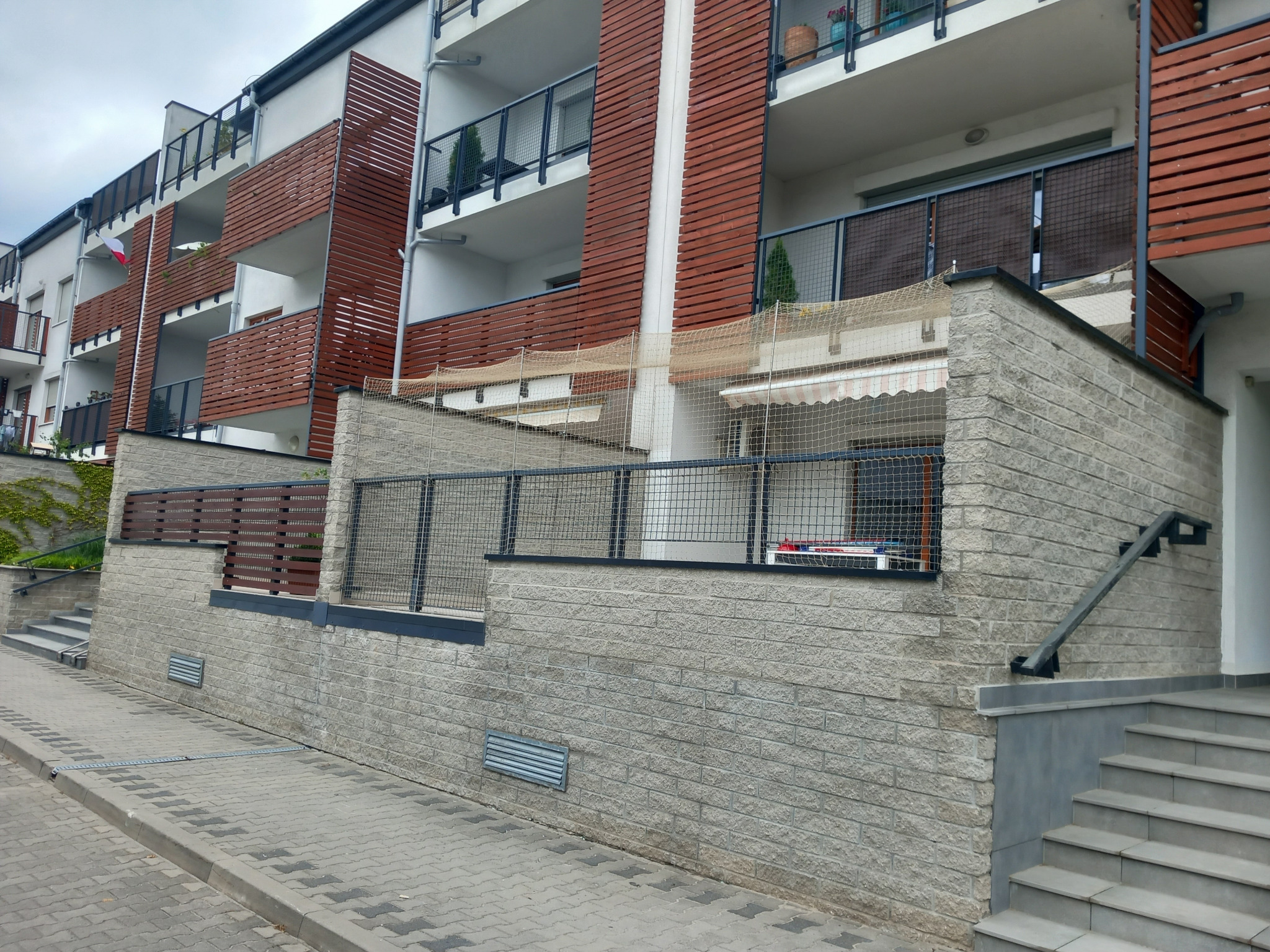Wielopiętrowy budynek z balkonami zabezpieczonymi siatką, prawdopodobnie dla kota, widok z poziomu chodnika.