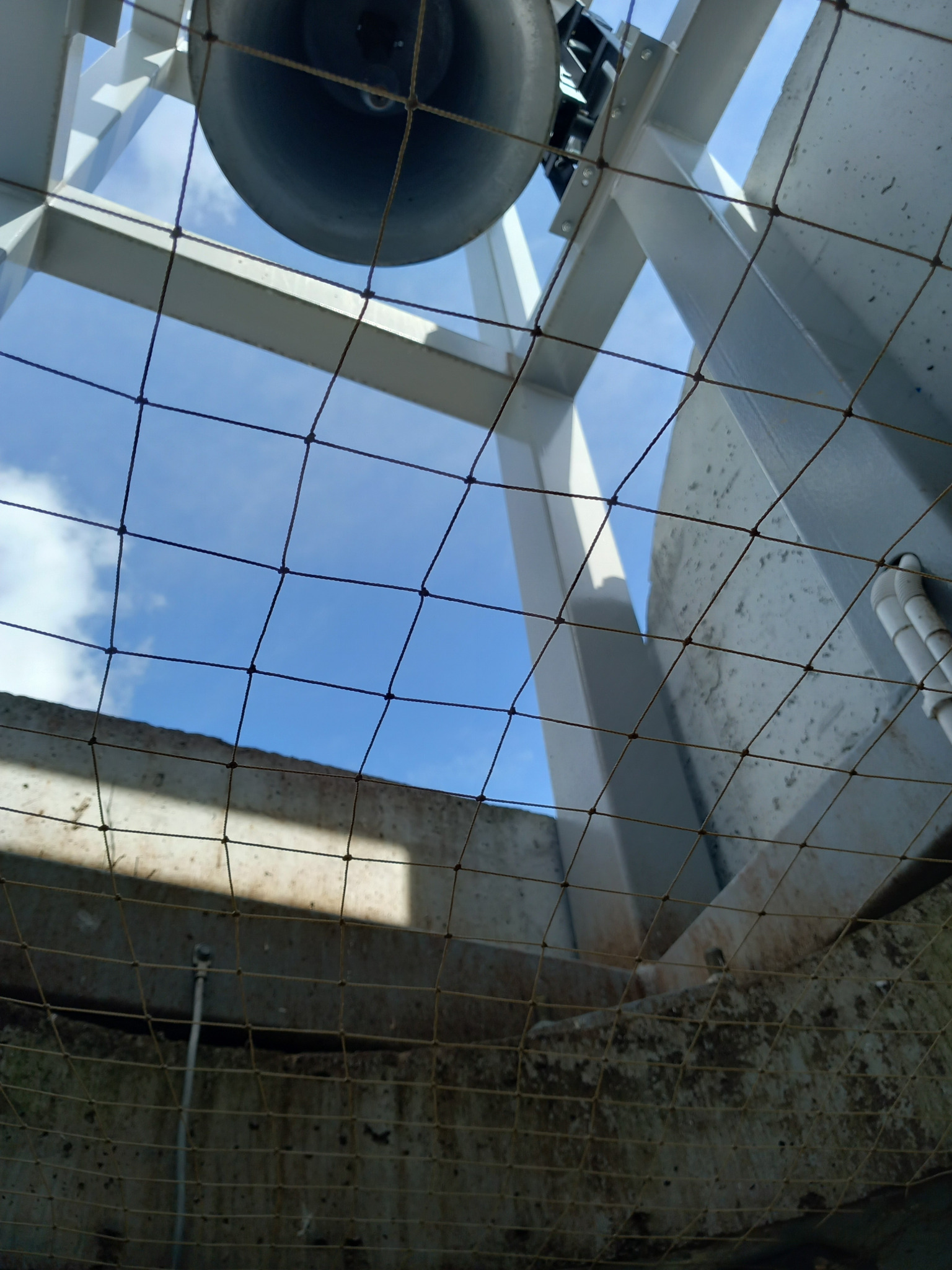 Ochronna siatka z drobnymi oczkami, zamontowana na metalowej konstrukcji, widok z dołu na błękitne niebo z chmurami.