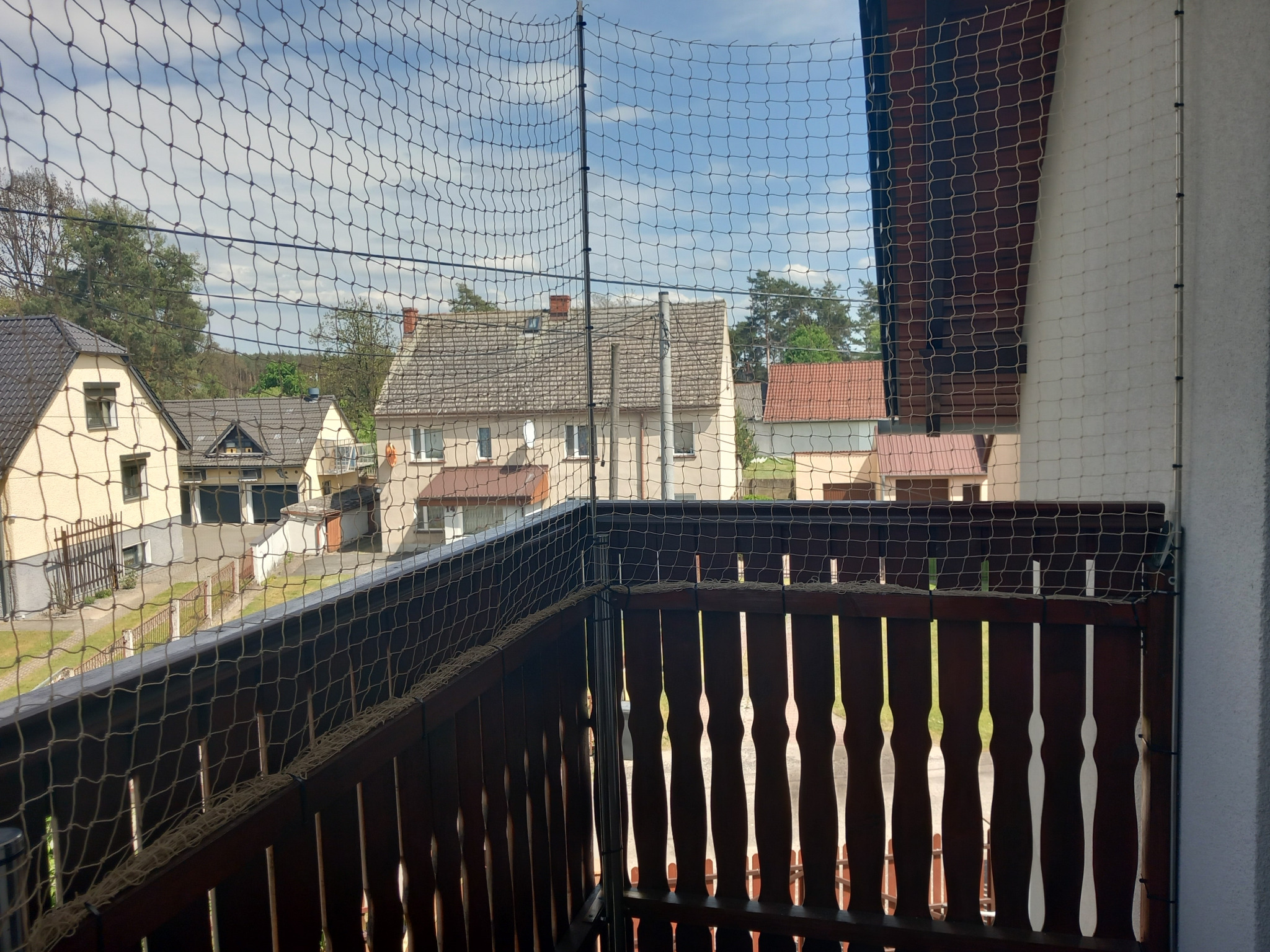 Ochronna siatka zamontowana na balkonie z drewnianą balustradą, widok na domy i drzewa w tle.