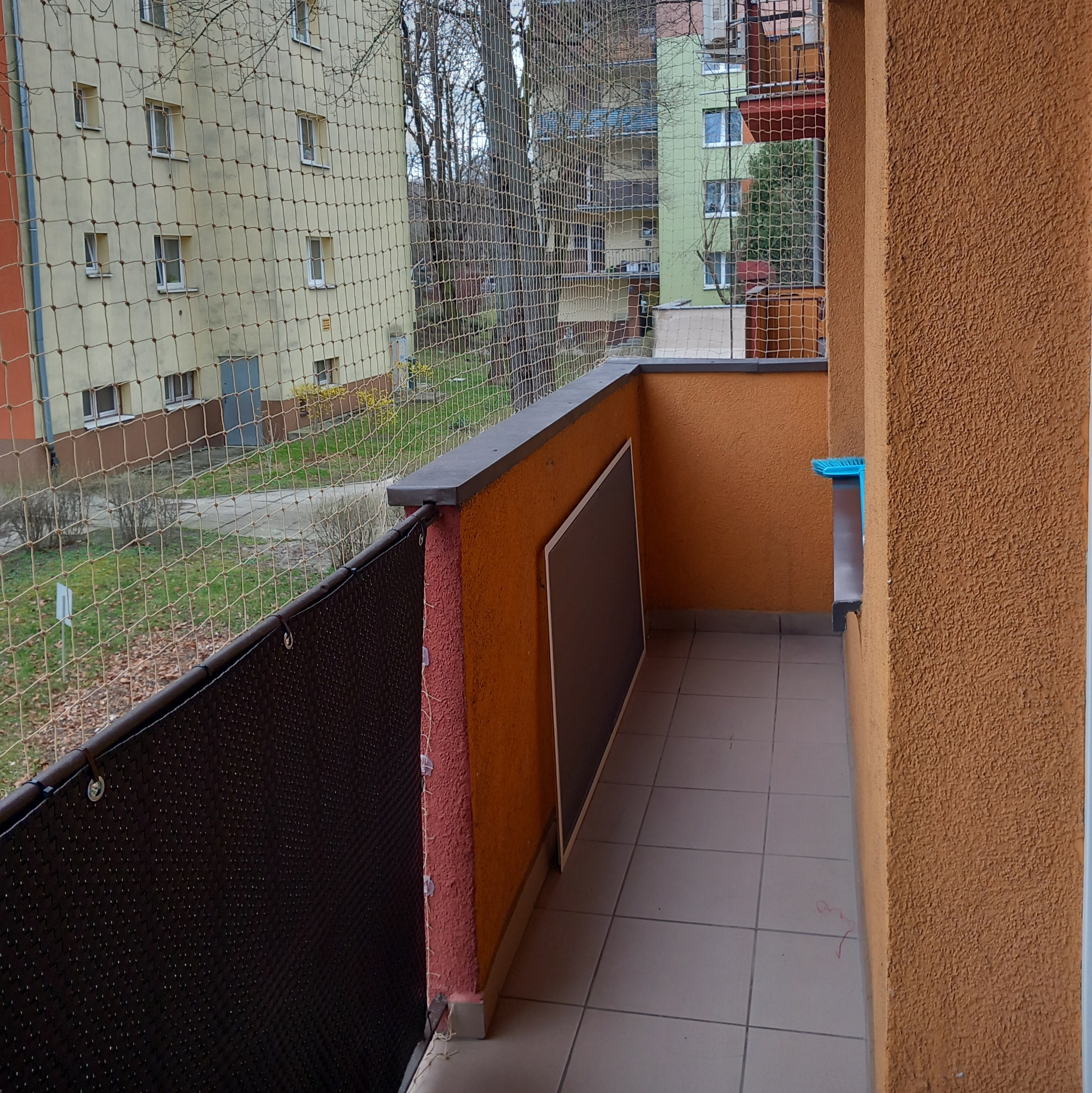 Zabezpieczony balkon siatką z drobnymi oczkami, widok na blok mieszkalny i zieleń, dodatkowo zamontowana czarna osłona balkonowa.