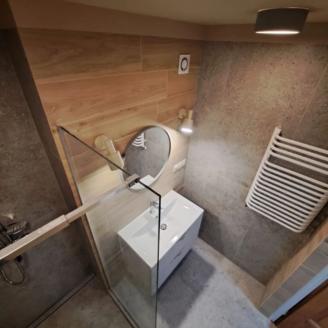 Nowoczesna łazienka po remoncie: kabina prysznicowa ze szklaną ścianką, umywalka z szafką, lustro owalne i grzejnik drabinkowy na tle płytek imitujących drewno i kamień.