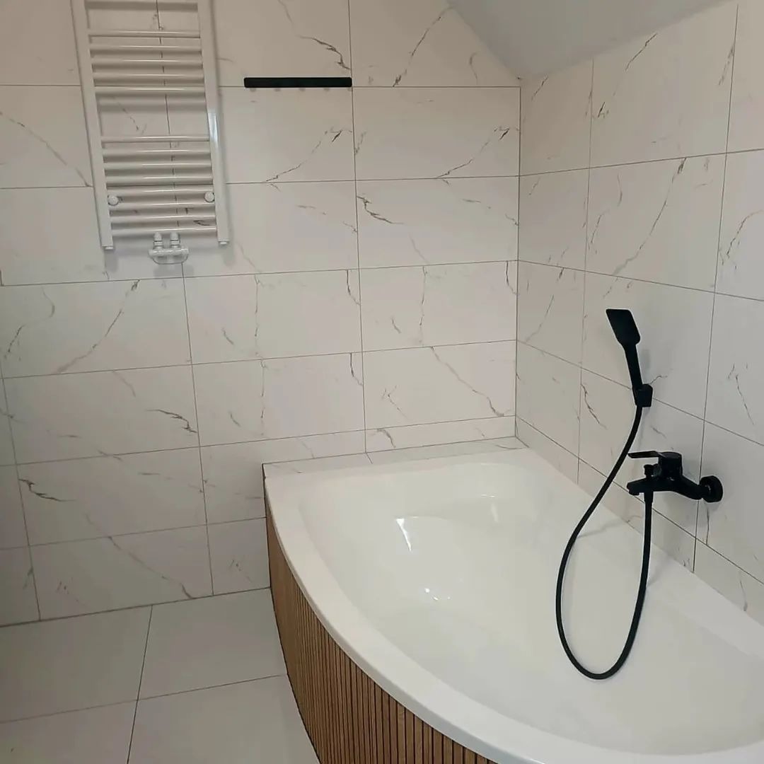 Biała wanna z czarną armaturą i marmurowe płytki w łazience. Grzejnik drabinkowy i drewniane wykończenie wanny dodają elegancji. Nowoczesny design.