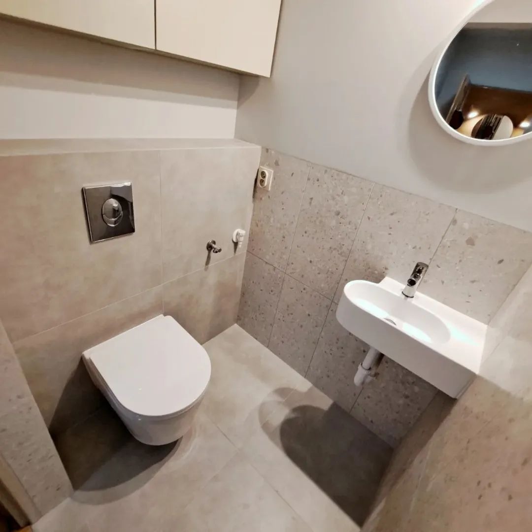 Nowoczesna, beżowa łazienka z podwieszaną toaletą, umywalką nablatową i okrągłym lustrem. Minimalistyczny design, płytki imitujące lastryko na ścianie.