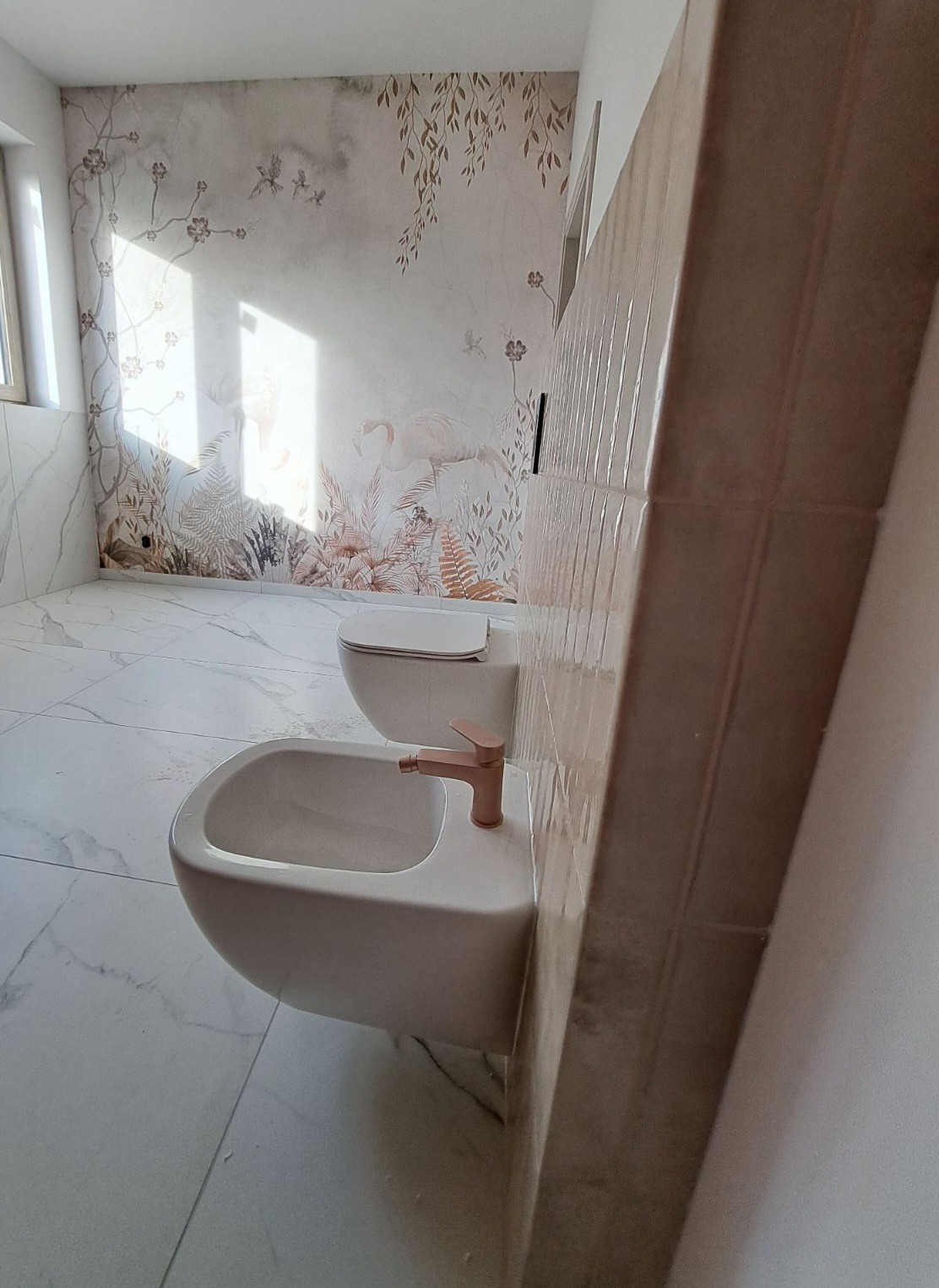 Jasna łazienka z marmurową podłogą i ścianami, bidet z miedzianą armaturą, toaleta i ściana z motywem roślinnym. Ukończone wykończenie wnętrza.