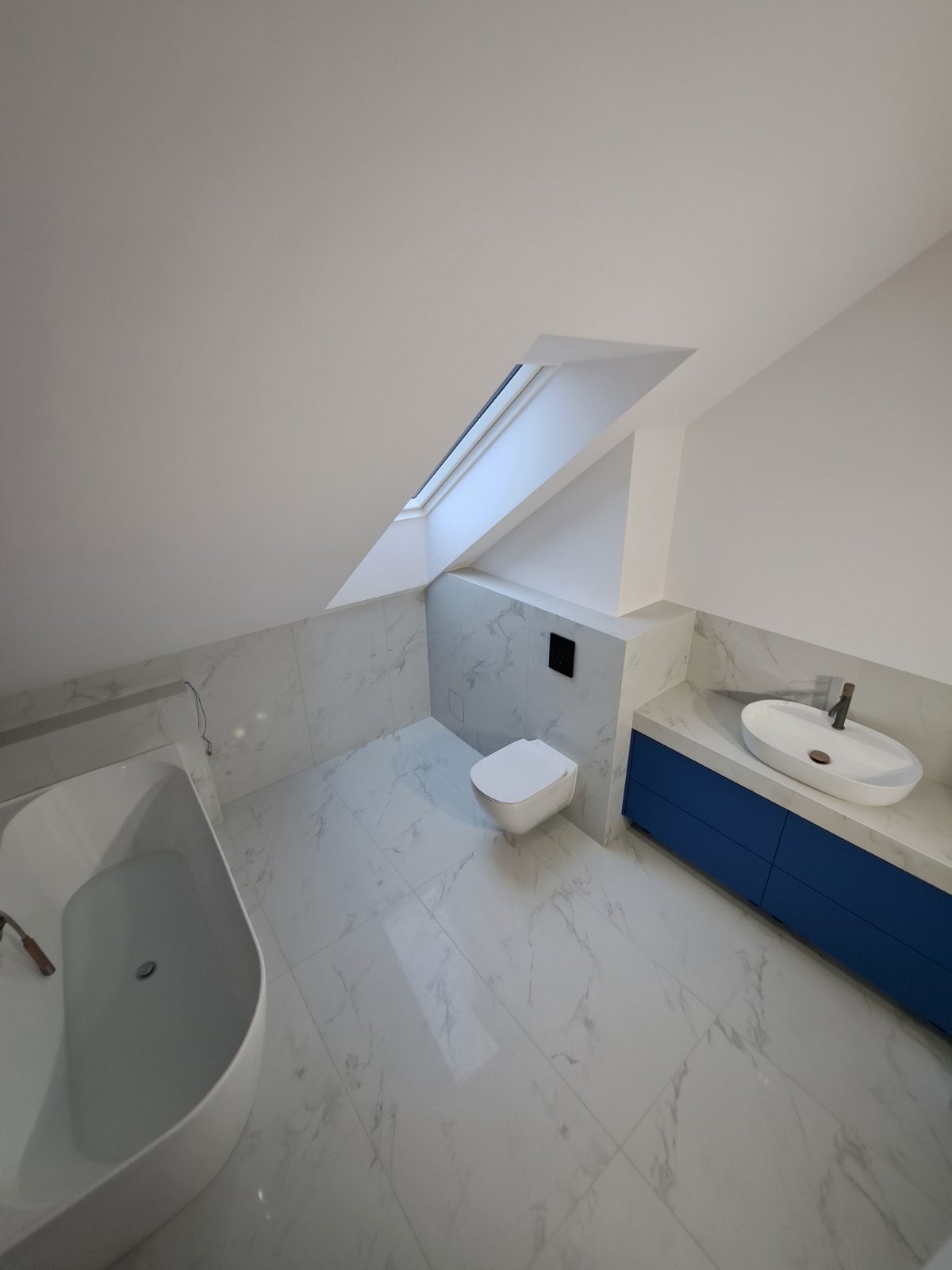 Jasna łazienka na poddaszu z marmurowymi płytkami, wolnostojącą wanną, toaletą podtynkową i szafką z umywalką w kolorze kobaltowym. Okno dachowe doświetla wnętrze.