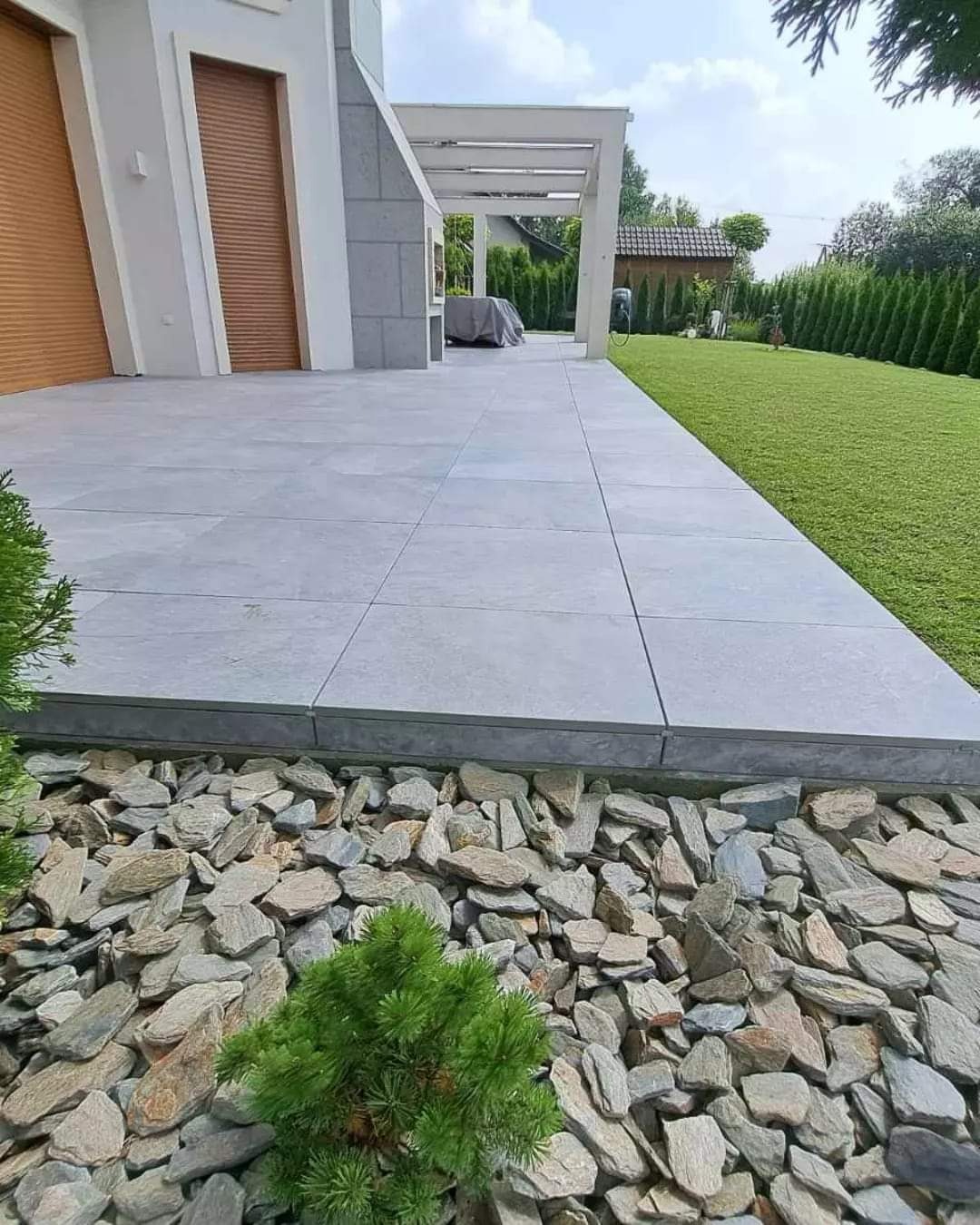 Nowoczesny taras z szarych płyt, obrzeże z kamieni ozdobnych i zielony trawnik. Widok na dom z roletami i pergolą. Minimalistyczny design.