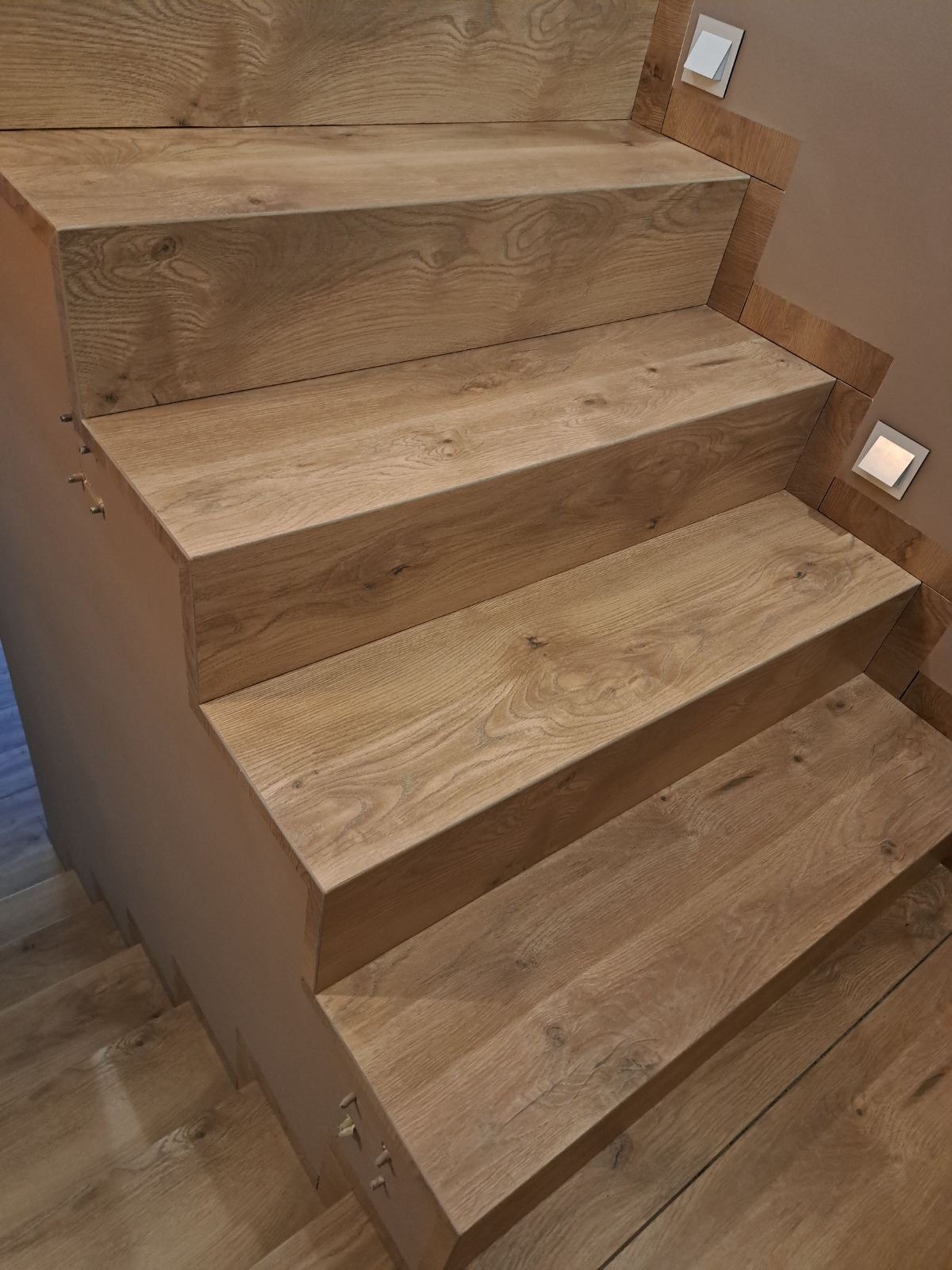 Drewniane schody z oświetleniem punktowym wzdłuż ściany, wykonane w Krakowie. Widoczne detale usłojenia drewna i precyzyjne wykończenie.