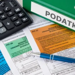 Kalkulator, długopis i formularze PIT obok zielonego segregatora z napisem 'Podatki'. Zestawienie narzędzi pracy doradcy podatkowego w bliskim ujęciu.