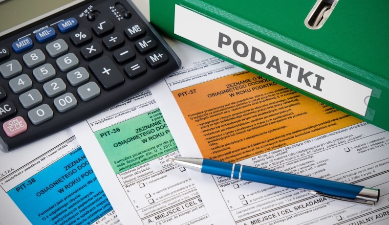 Kalkulator, długopis i formularze PIT obok zielonego segregatora z napisem 'Podatki'. Zestawienie narzędzi pracy doradcy podatkowego w bliskim ujęciu.
