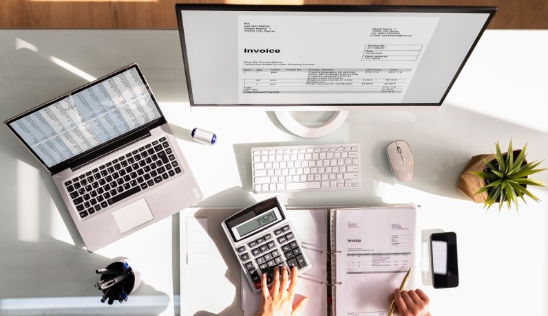 Biurko z laptopem, monitorem prezentującym fakturę, kalkulatorem w użyciu, dokumentami i akcesoriami biurowymi w jasnym, nowoczesnym stylu.