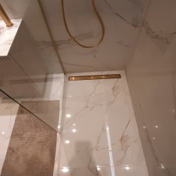 Rem & bud - Nowoczesna kabina prysznicowa z marmurowymi płytkami, złotą armaturą i odpływem liniowym, widok z góry z fragmentem osoby w kadrze.