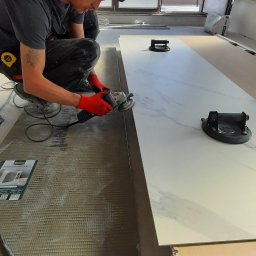 Rem & bud - Pracownik w czerwonych rękawicach szlifuje krawędź płytki gresowej o wzorze marmuru szlifierką kątową, obok leży płyta budowlana do montażu pod płytki, widoczne przyssawki do przenoszenia płytek.