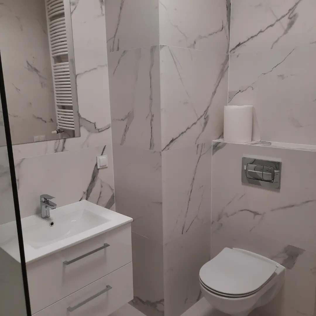 Nowoczesna łazienka z białymi płytkami imitującymi marmur, wisząca toaleta z chromowanym przyciskiem spłukiwania, umywalka z szafką i grzejnik drabinkowy.