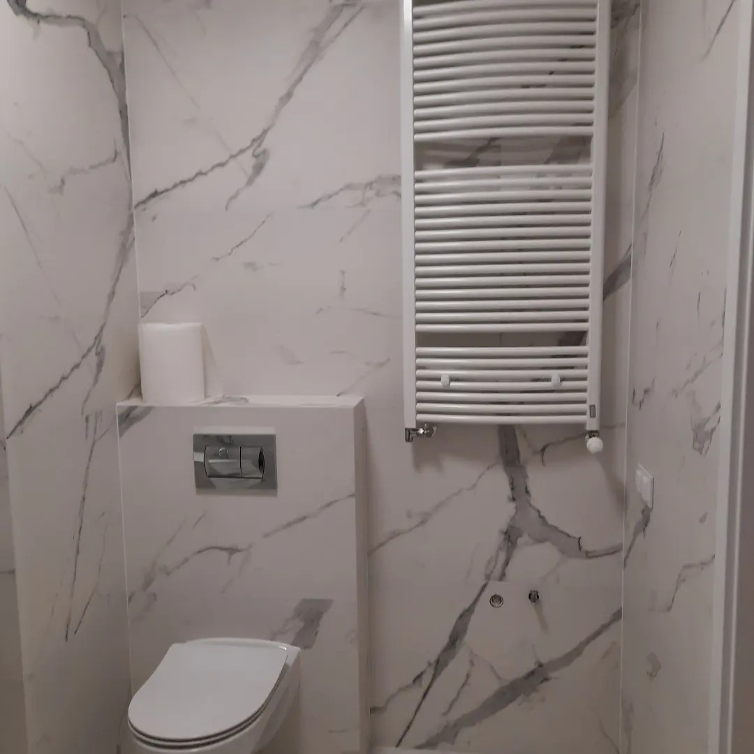 Biała toaleta podwieszana z chromowanym przyciskiem spłukującym, biały grzejnik drabinkowy i ściany wykończone płytkami imitującymi biały marmur z szarymi żyłami.