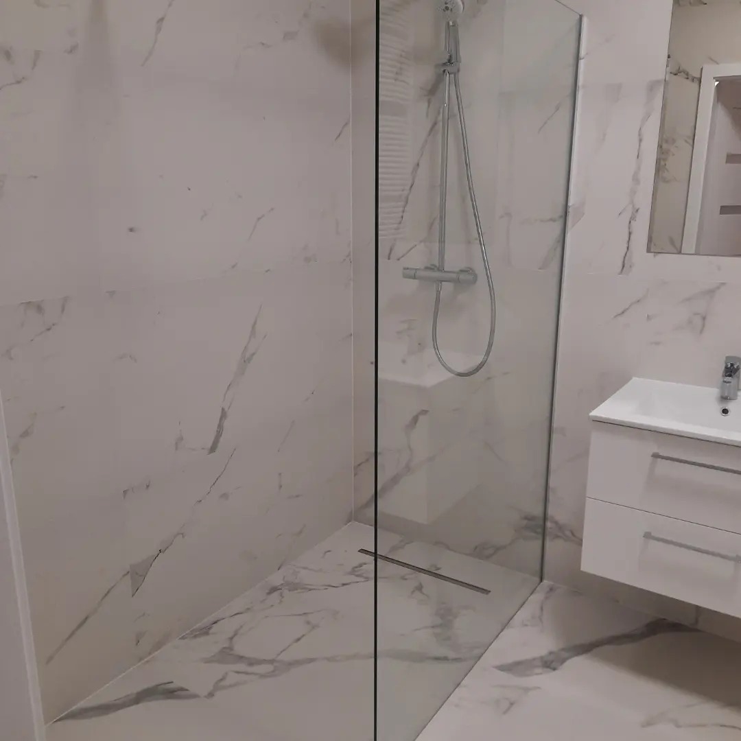 Nowoczesna kabina prysznicowa typu walk-in z odpływem liniowym, wykończona białymi płytkami imitującymi marmur, z umywalką na szafce w tle.